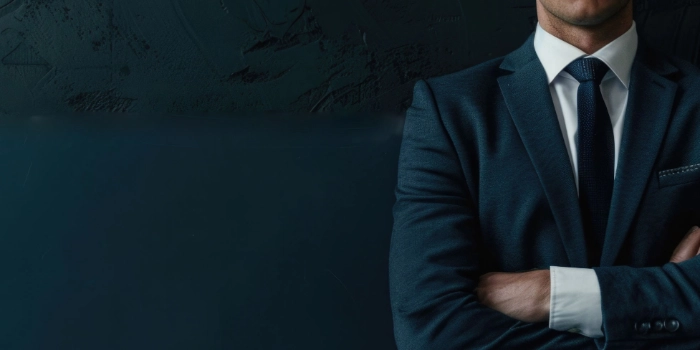 Professional consultant in dark suit with arms crossed in front of textured wall – symbolic header image for Advisory & Consulting Services in the field of anti-financial crime compliance.